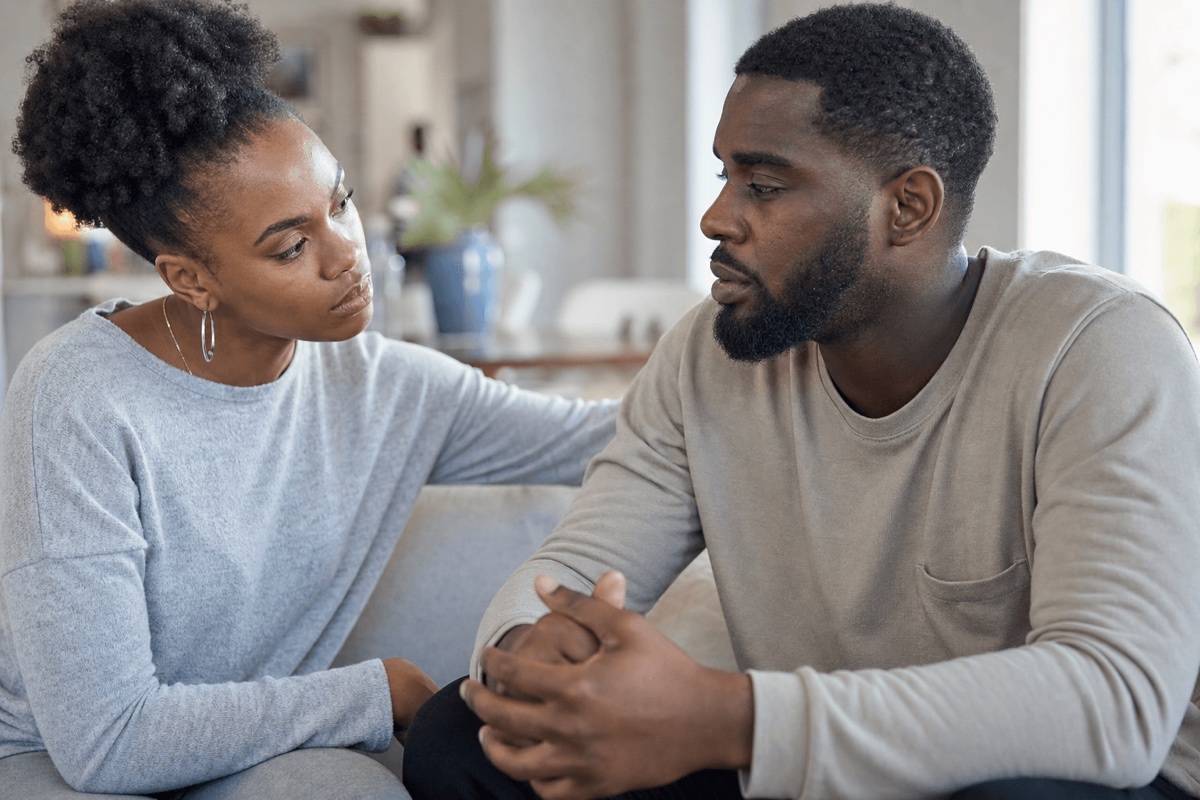 Two adults sitting close on a couch in calm conversation, one listening with concern