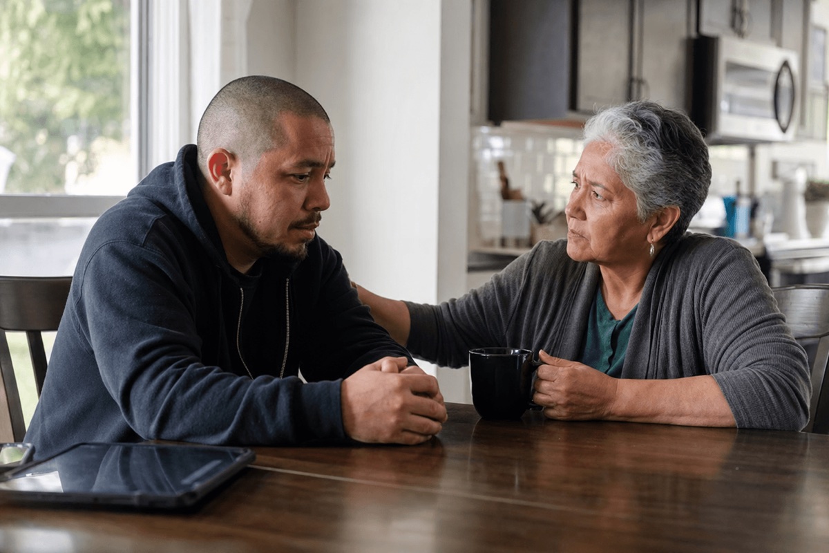 A worried adult sitting with an older family member at a kitchen table during a serious, supportive conversation