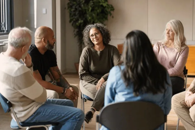 A small support group sitting in a circle of chairs in a bright community room during a recovery meeting