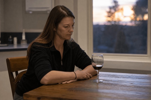 A person sitting alone at a kitchen table at dusk, looking thoughtfully at a glass of wine