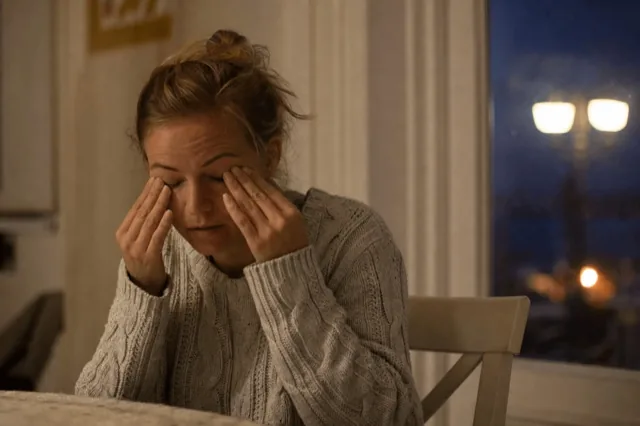 A person rubbing tired eyes in soft evening light, with blurred lamplight behind them