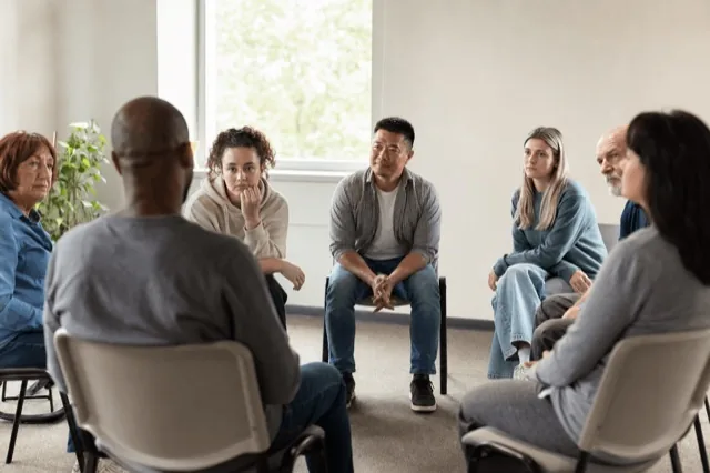 A small addiction-treatment group therapy session in a bright, calm room with people seated in a circle