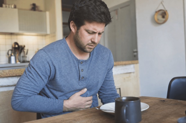 A person holding their upper abdomen with a discomforted expression at a kitchen table