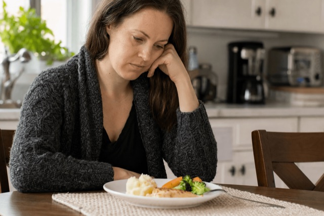 A person sitting at a kitchen table looking thoughtfully at a plate of food in soft morning light