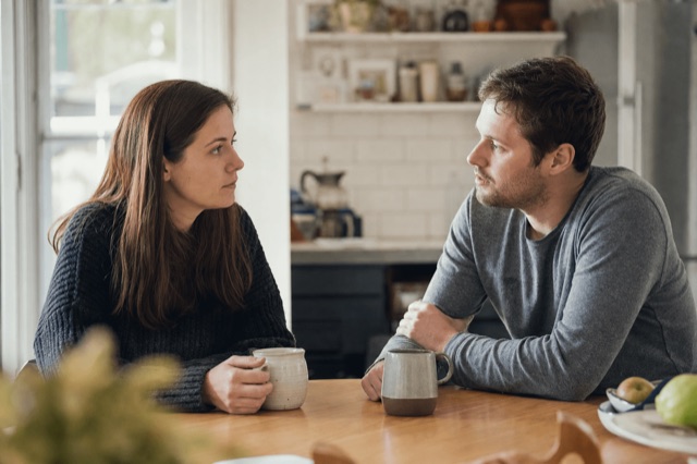 A couple sitting together at a kitchen table having a calm, serious conversation in soft morning light