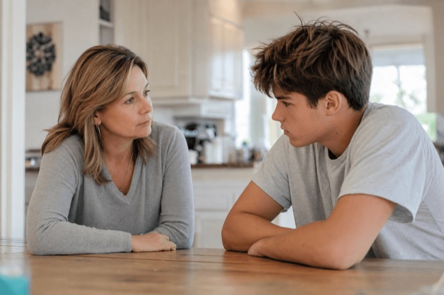 A parent and teenager talking together at a kitchen table in soft daylight