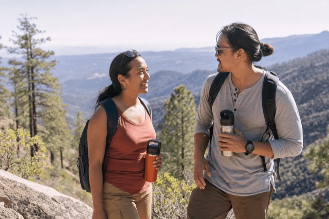 Two friends hiking on a sunlit mountain trail with water bottles, enjoying an alcohol-free getaway