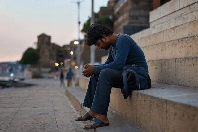 A person sitting alone on city steps at dusk, bag beside them, looking down in quiet difficulty