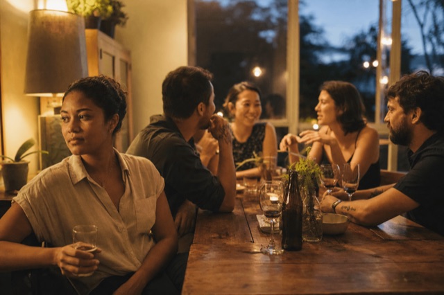A small group of friends holding drinks at a relaxed evening gathering, with one person looking quietly reflective